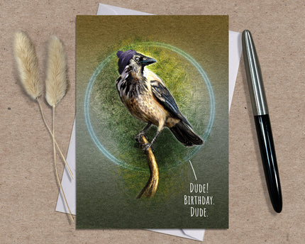 Greeting card with a bird illustration and text 'Dude! Birthday, Dude.' on a brown surface with a pen and dried plants.