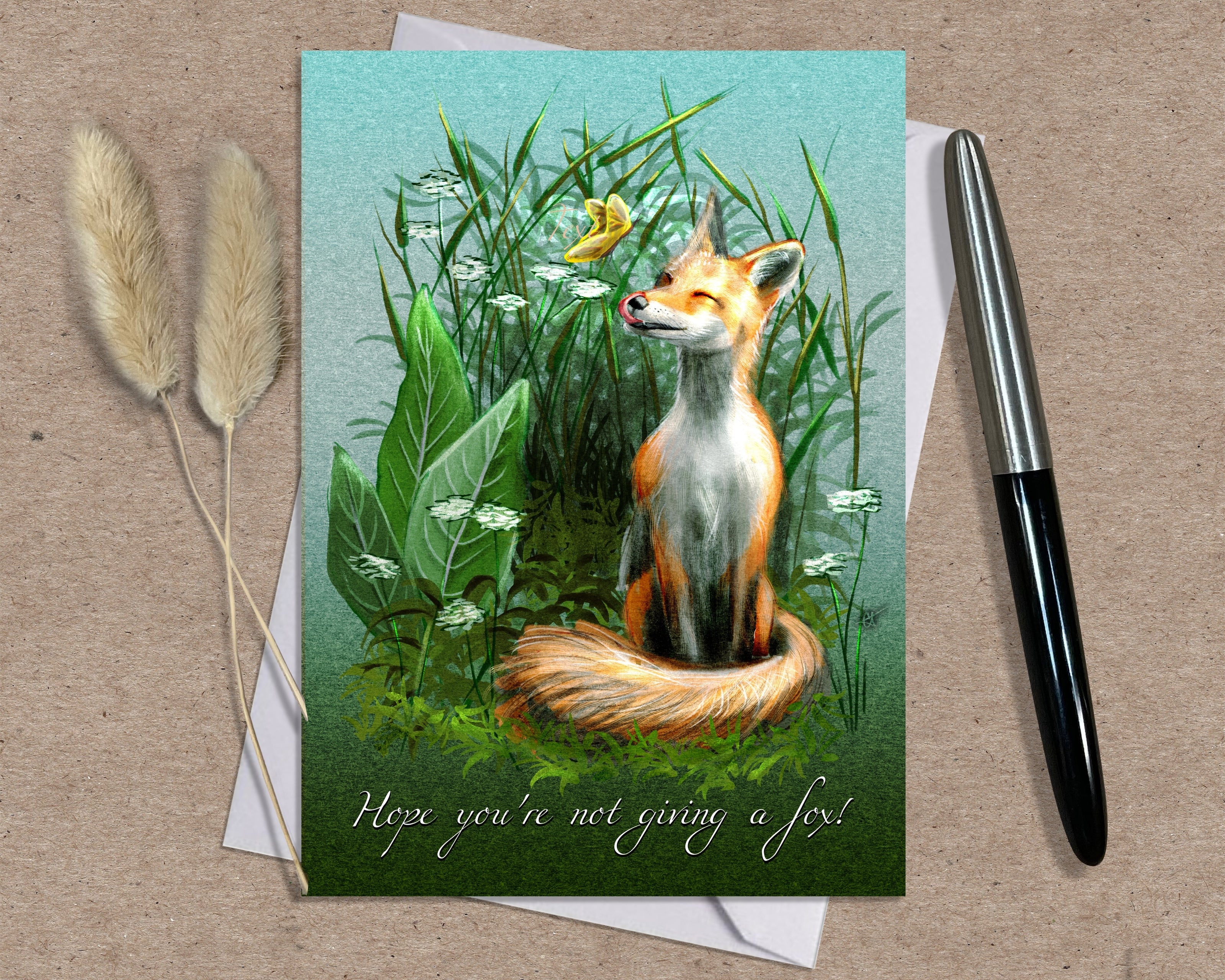 Greeting card with a fox illustration on a grassy background, accompanied by a pen and dried plants. Text says, 