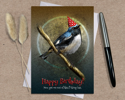 Birthday card with a bird wearing a party hat on a branch, placed on a textured surface with a pen and dried plants.