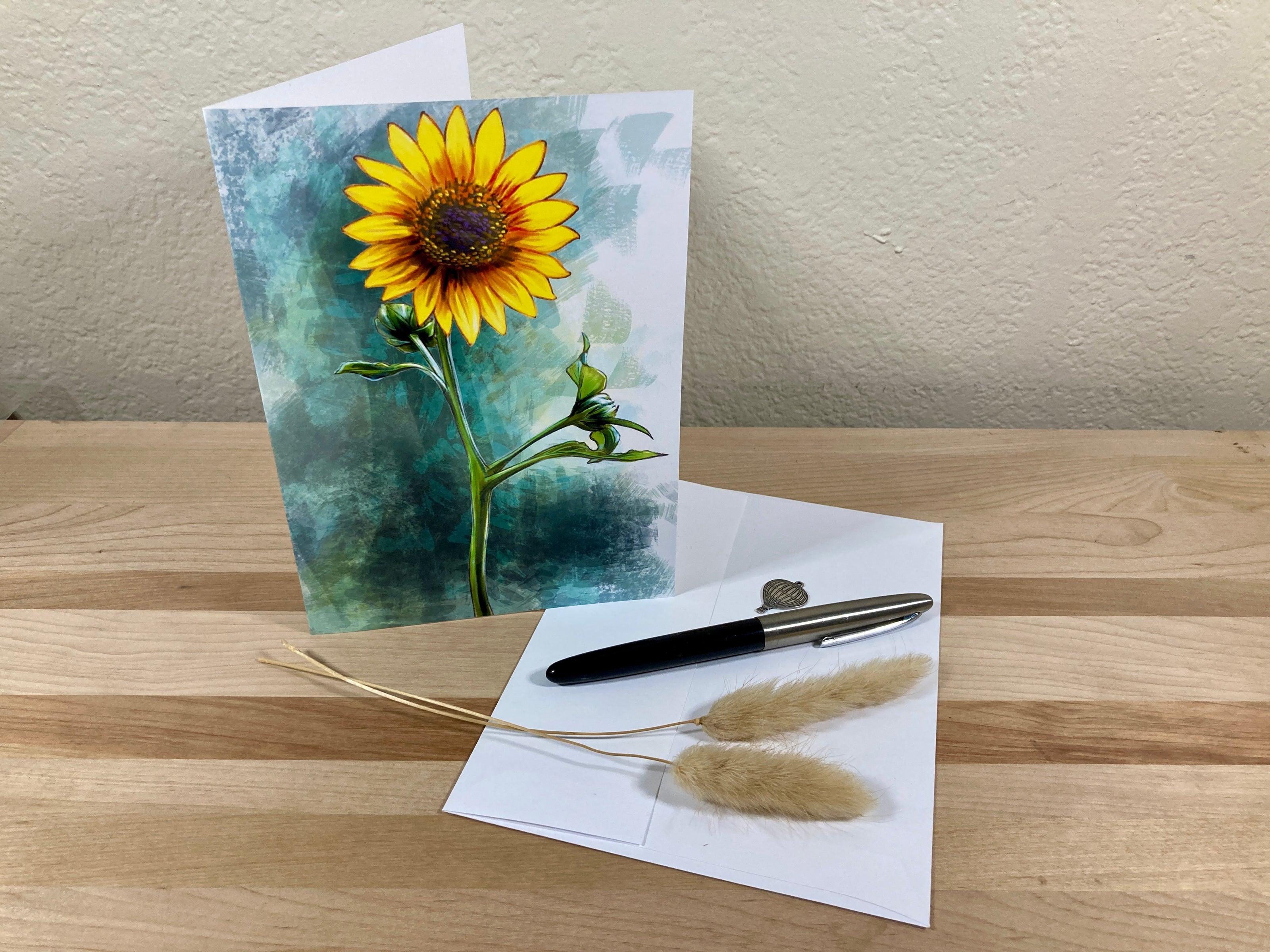 Greeting card with sunflower design on a wooden surface with a pen and dried plants.