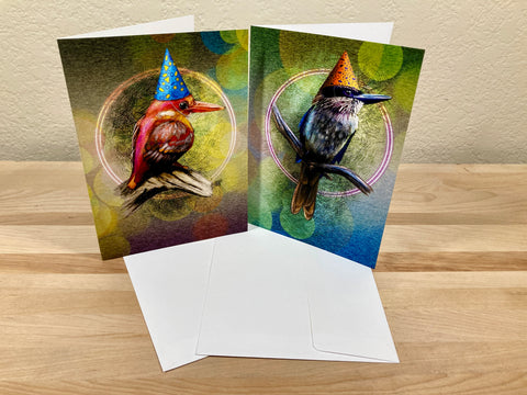 Two greeting cards with bird illustrations wearing party hats on a wooden surface.