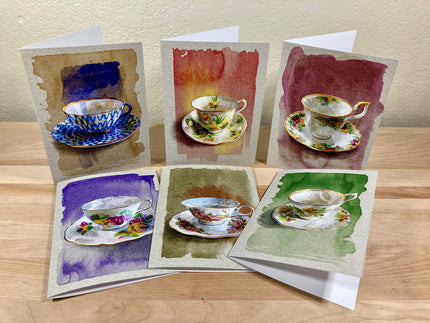 Set of six greeting cards with watercolor designs of teacups and saucers on a wooden surface.