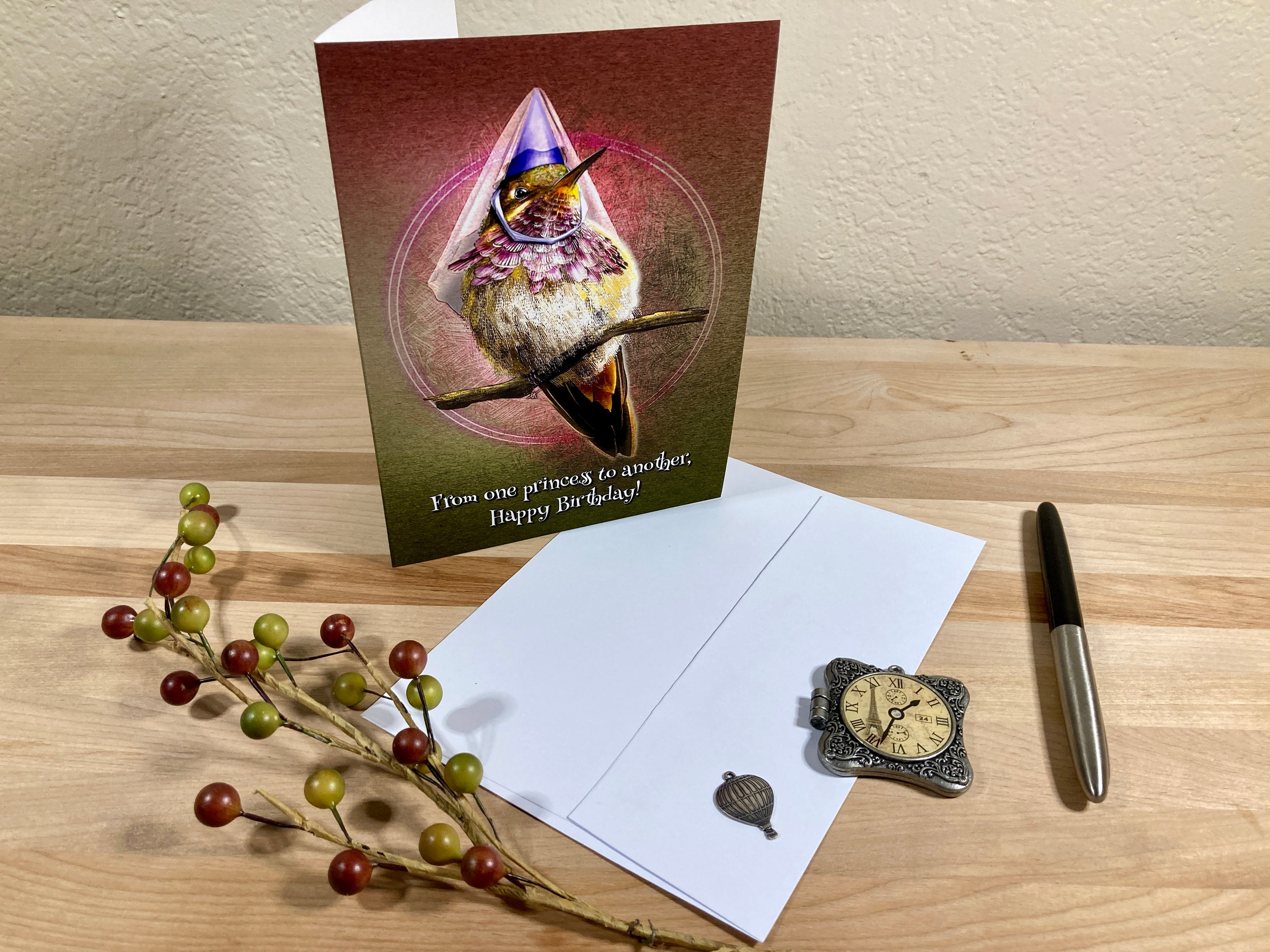 Greeting card with a hummingbird design on a wooden surface with a pen and decorative branch.