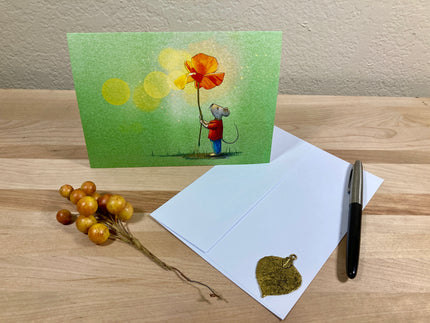 Greeting card with a mouse holding a flower, next to an open envelope and pen on a wooden surface.