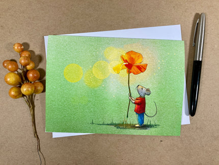 Greeting card with illustration of a mouse holding a flower, placed on a brown surface with a pen and berries.
