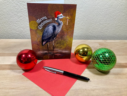 Christmas card with heron wearing a Santa hat, surrounded by festive balls and a pen on a wooden surface.