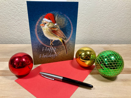 Christmas card with a bird wearing a Santa hat, surrounded by festive balls and a pen on a wooden surface.