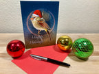Christmas card with a bird wearing a Santa hat, surrounded by festive balls and a pen on a wooden surface.