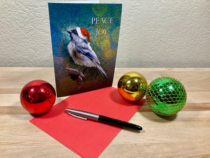 Christmas card with a bird illustration, red envelope, pen, and colorful ornaments on a wooden surface.
