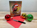 Happy Holidays card with a bird illustration, red pen, and Christmas ornaments on a wooden surface.