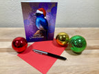Christmas card with a bird design, pen, red paper, and colorful ornaments on a wooden surface.