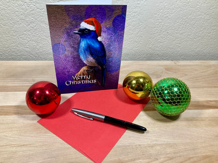 Christmas card with a bird design, pen, red paper, and colorful ornaments on a wooden surface.