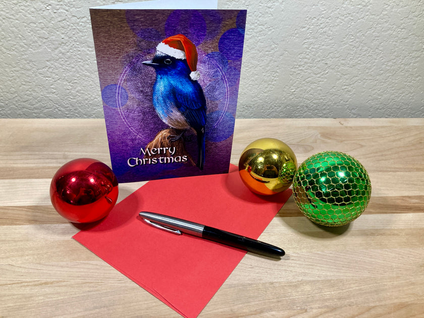 Christmas card with a bird design, pen, red paper, and colorful ornaments on a wooden surface.
