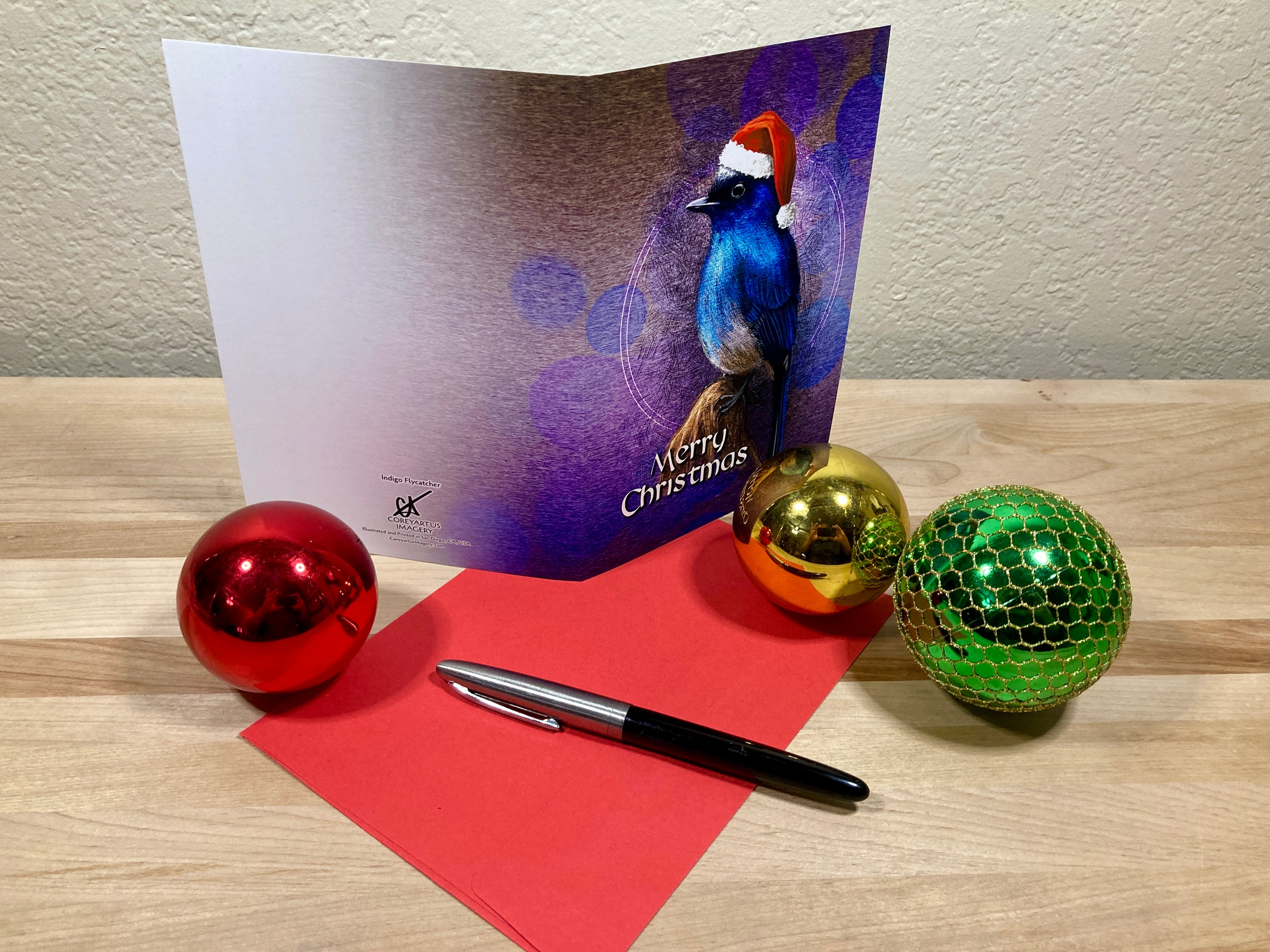 Christmas card with a bird illustration, pen, and colorful ornaments on a wooden surface.
