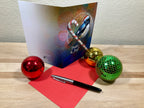 Greeting card with candy cane and bird design, surrounded by Christmas ornaments on a wooden surface.