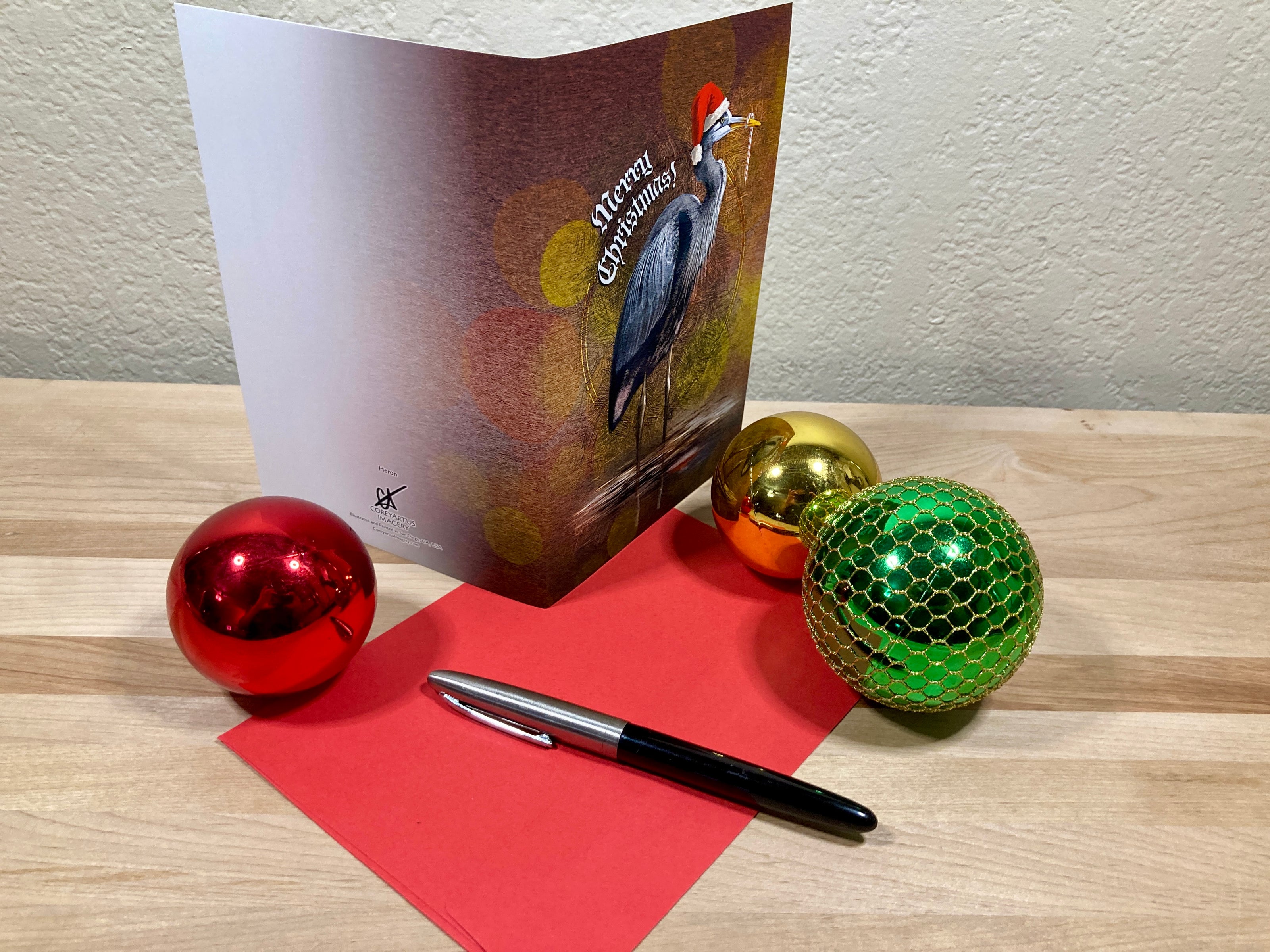 Christmas card with a bird design, red paper, pen, and colorful ornaments on a wooden surface.