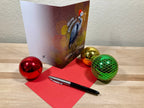 Christmas card with a bird design, red paper, pen, and colorful ornaments on a wooden surface.