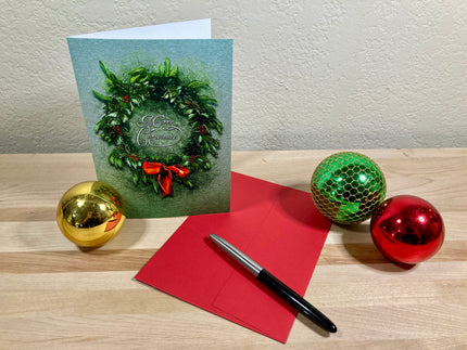 Christmas card with wreath design, red envelope, pen, and decorative balls on a wooden surface.