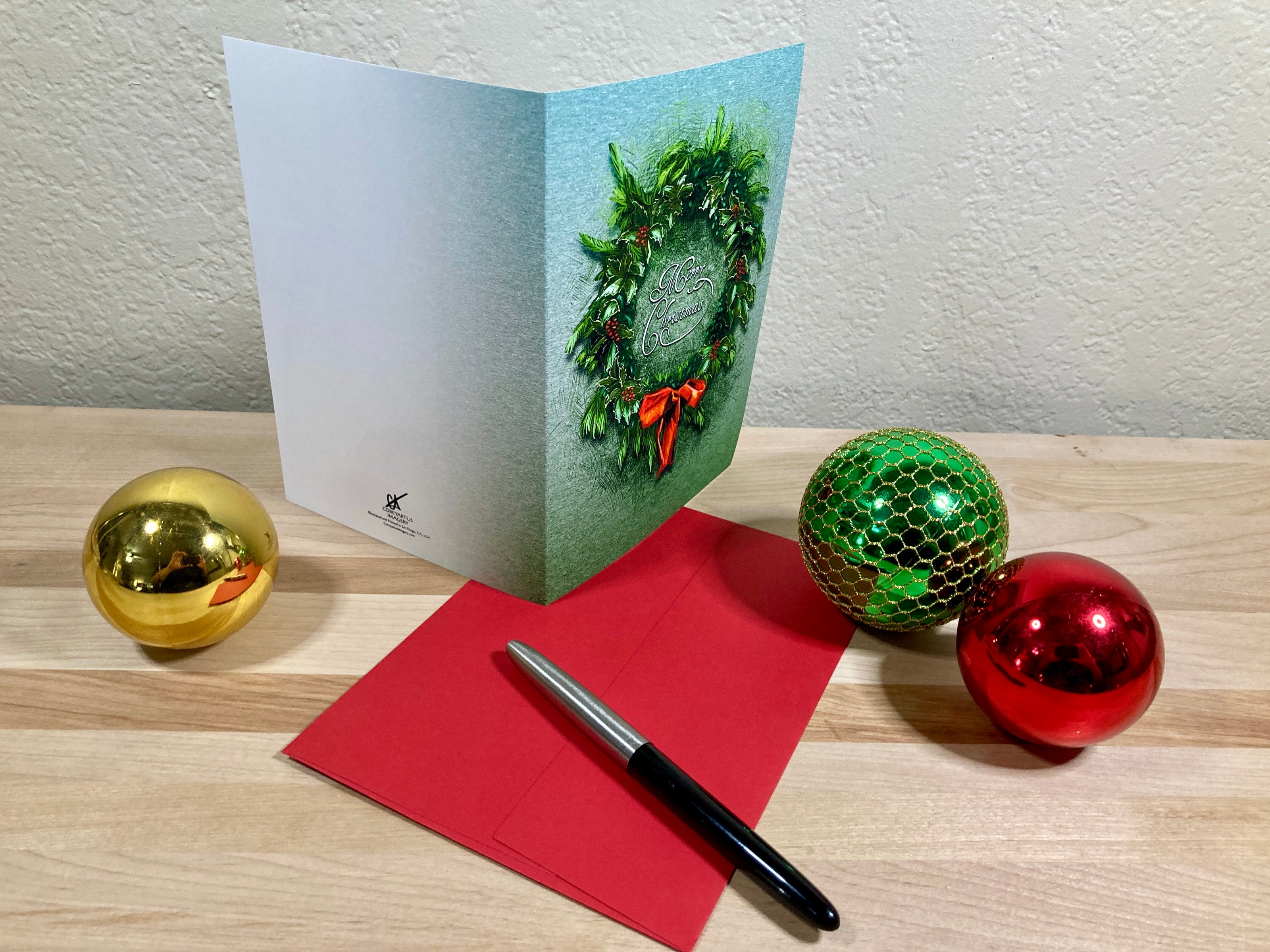 Christmas card with wreath design, red envelope, pen, and decorative balls on a wooden surface.