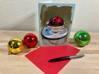 Greeting card with a teacup design, red ball, gold and silver pen, and Christmas ornaments on a wooden surface.