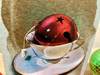 Red bell with a star on top inside a teacup on a saucer with a green decorative element.