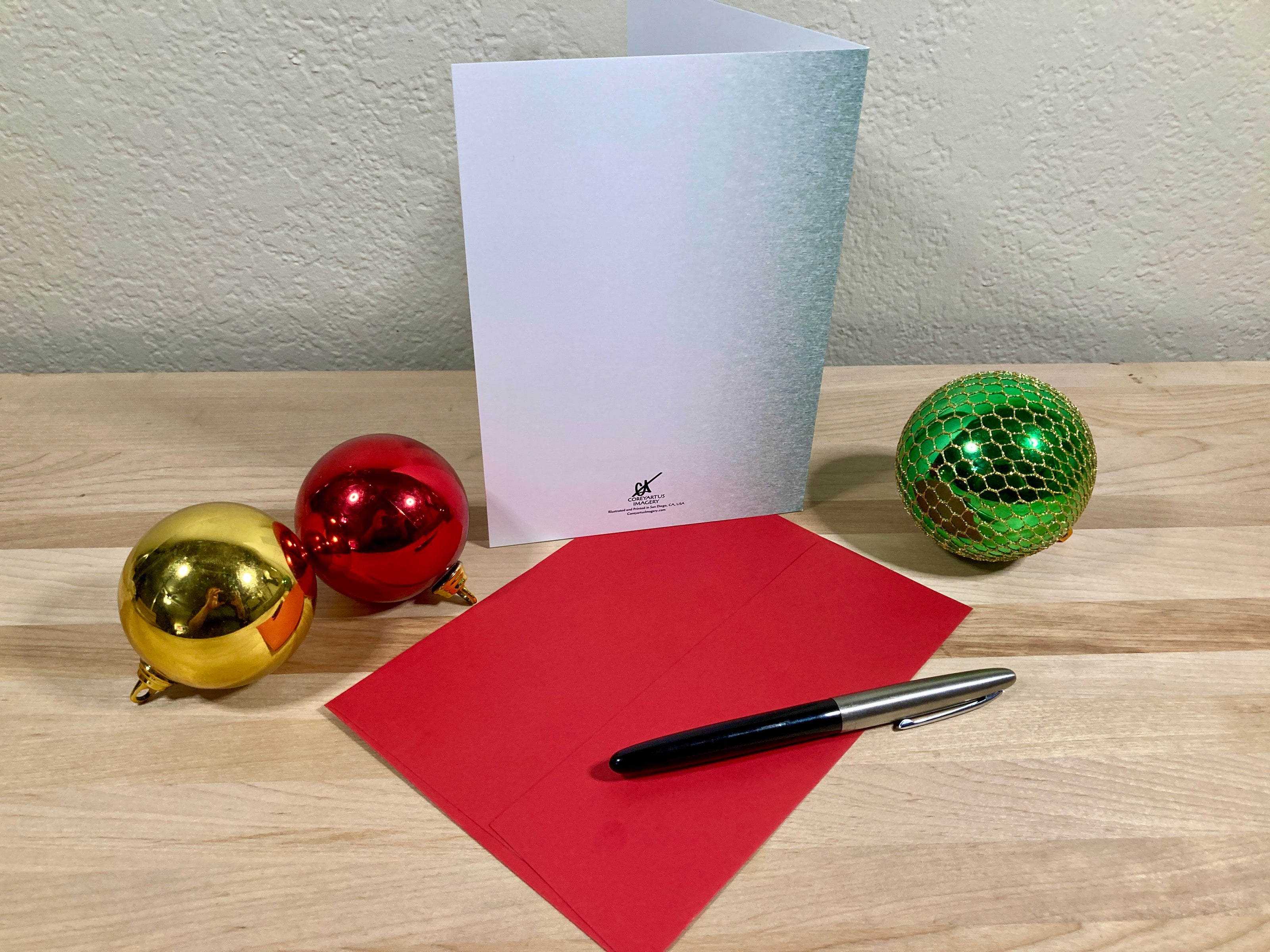 Greeting card, red envelope, pen, and Christmas ornaments on a wooden surface.