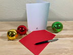 Greeting card, red envelope, pen, and Christmas ornaments on a wooden surface.