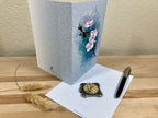 Floral card on a wooden surface with a pen and decorative clock.