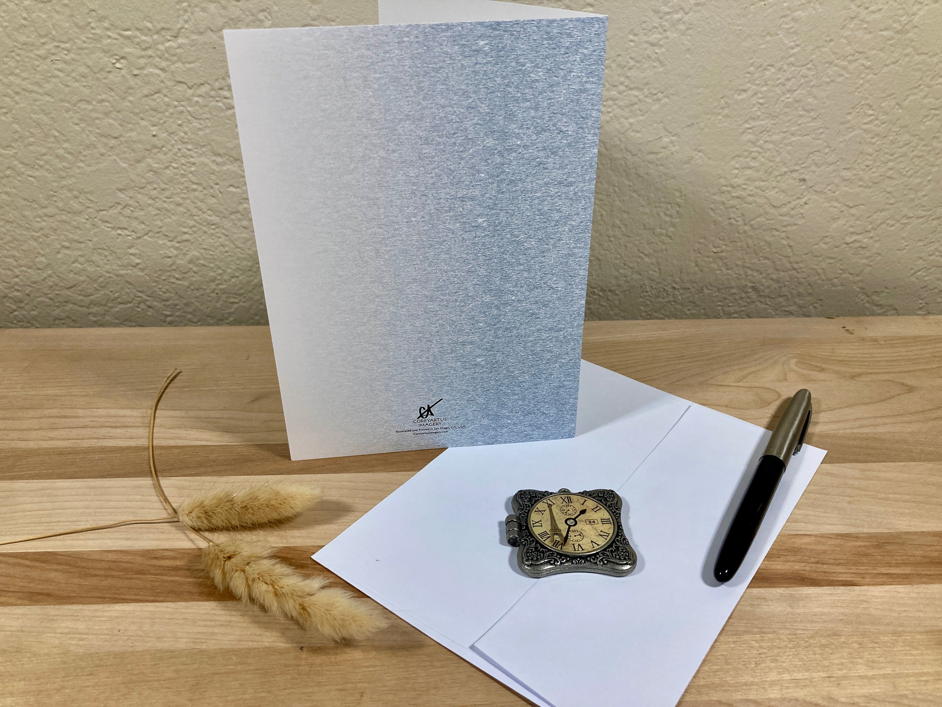 Back of a greeting card, pen, and decorative clock on a wooden surface with a neutral background