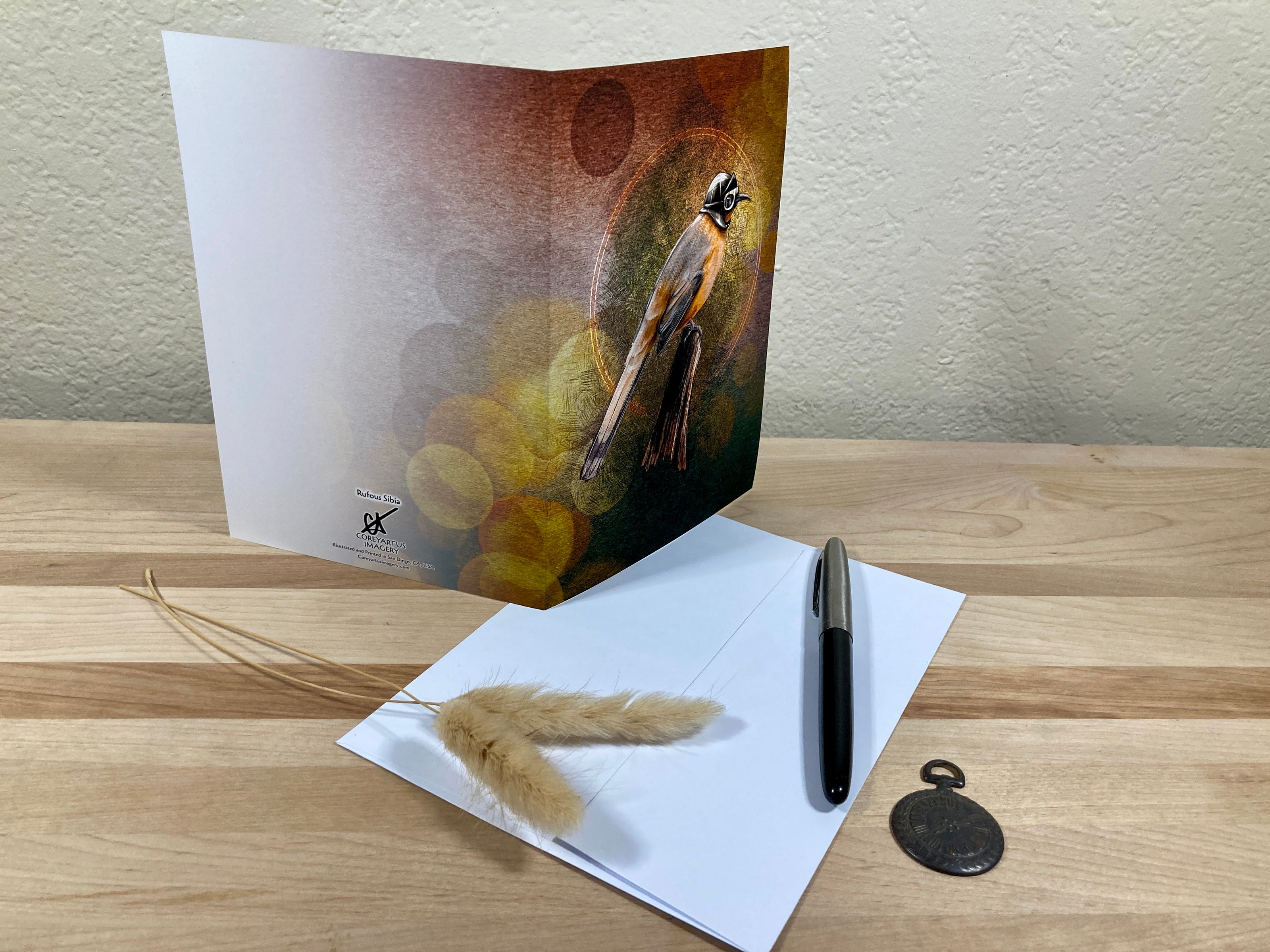 Greeting card with a bird design on a wooden surface with feathers and a pen.