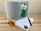 Greeting card with daffodil design, envelope, pen, and decorative stones on a wooden surface.
