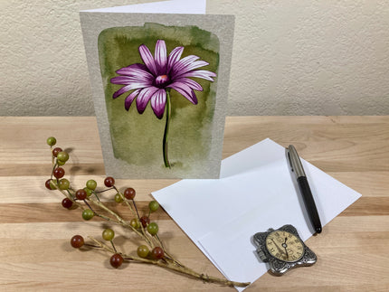 Greeting card with a purple flower illustration on a wooden surface with stationery items.
