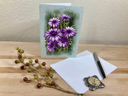 Greeting card with purple flowers on a wooden surface with a pen and clock.