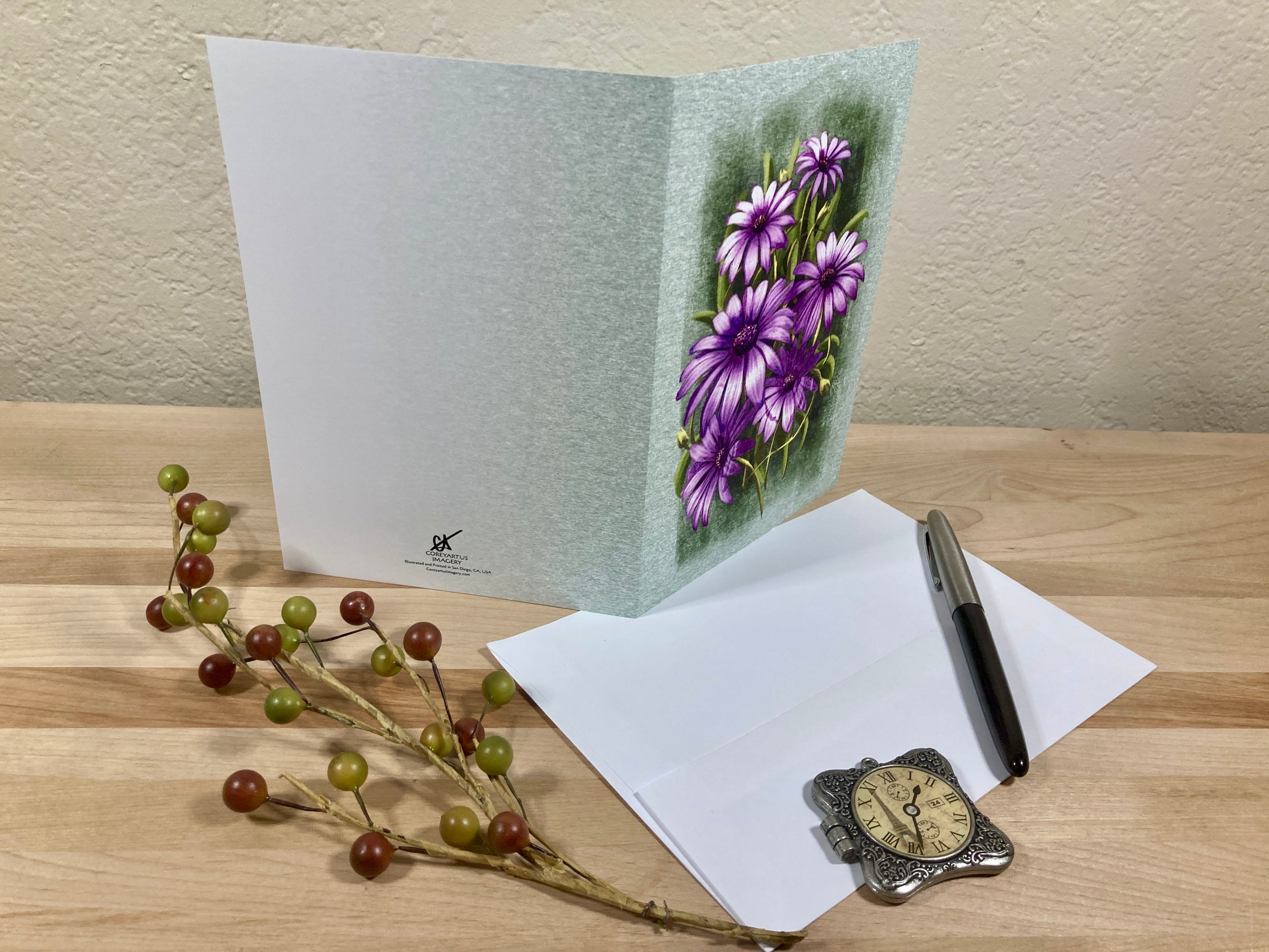 Greeting card with purple flowers, pen, watch, and decorative branch on a wooden surface.