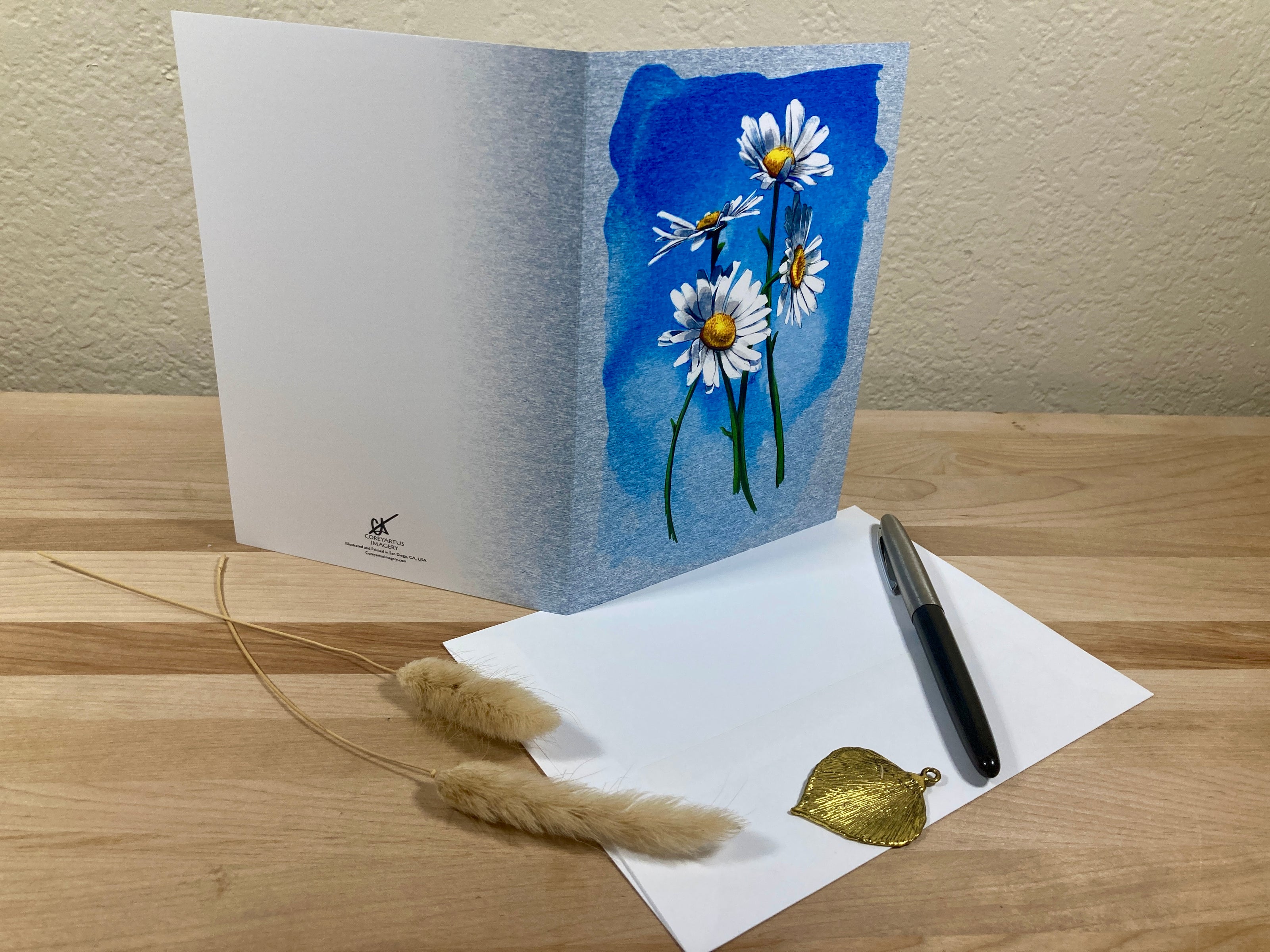 Greeting card with floral design on a wooden surface with a pen and decorative leaves.