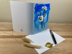 Greeting card with floral design on a wooden surface with a pen and decorative leaves.