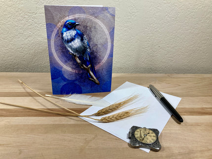 Greeting card with a blue bird design on a wooden surface with paper, pen, and decorative item.