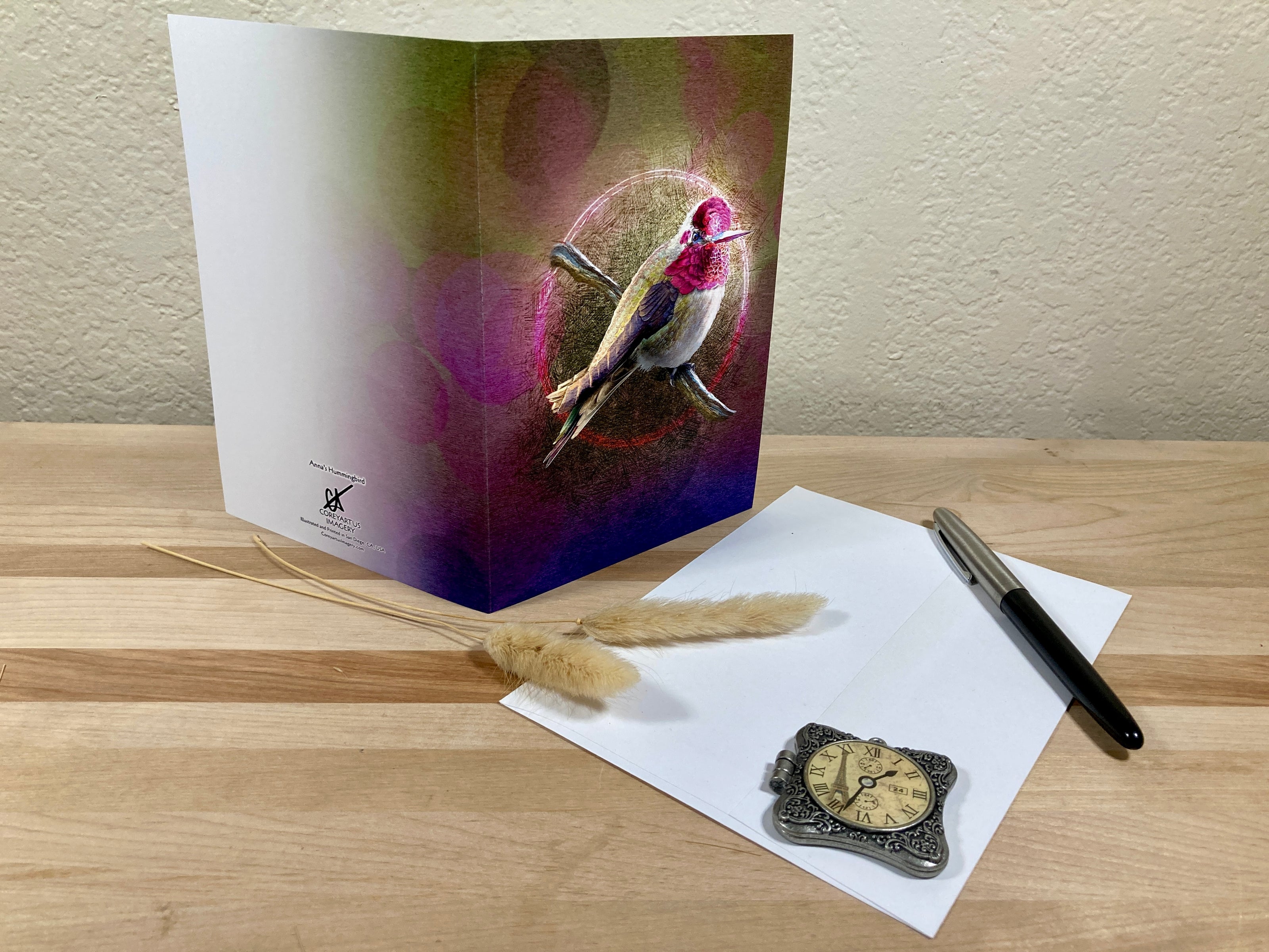 Greeting card with bird design, pen, paper, and pocket watch on a wooden surface.