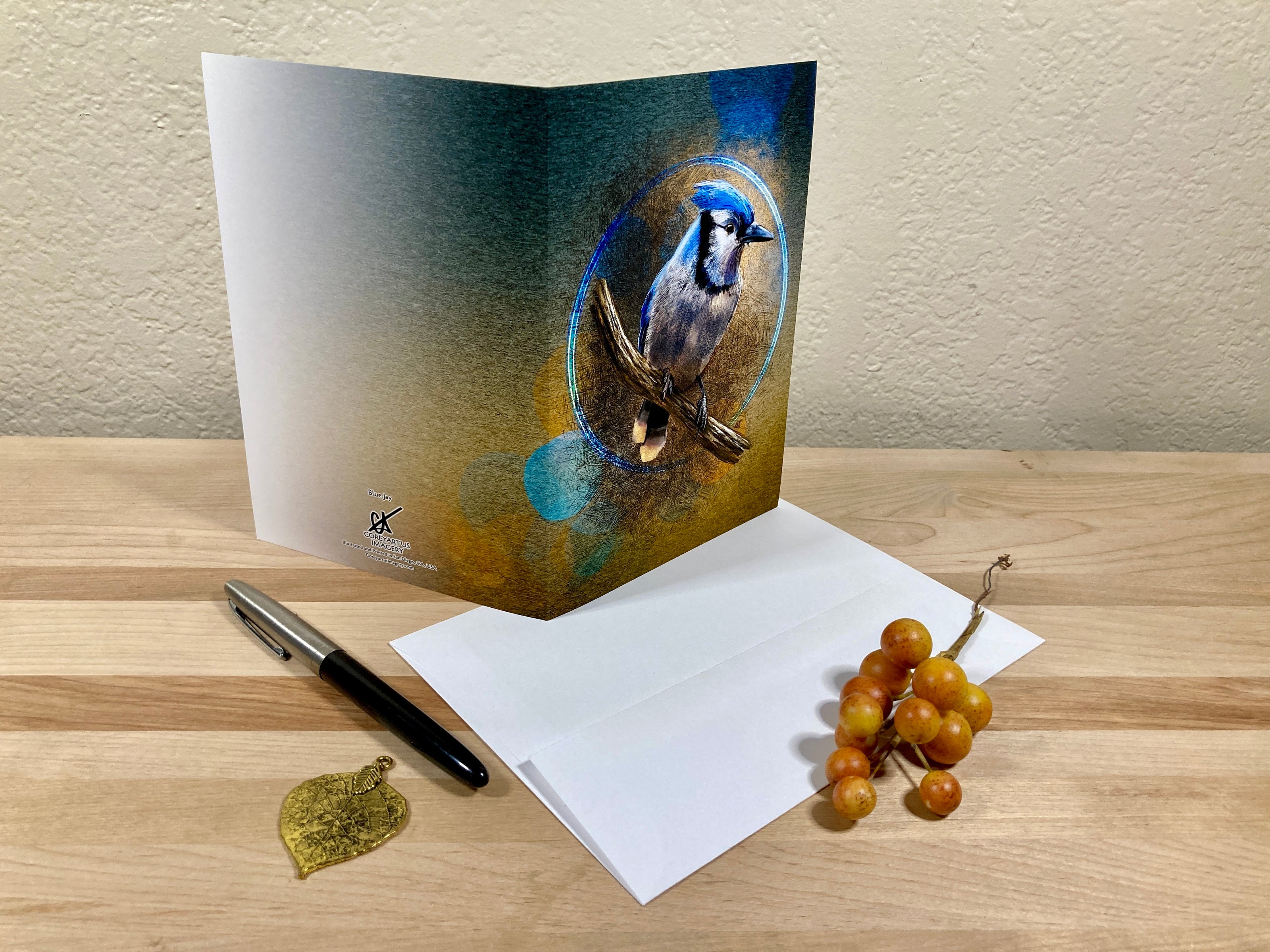 Greeting card with a bluejay design on a wooden surface with a pen and small decorative items.