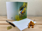 Greeting card with a bird illustration on a wooden surface with a pen and grapes.