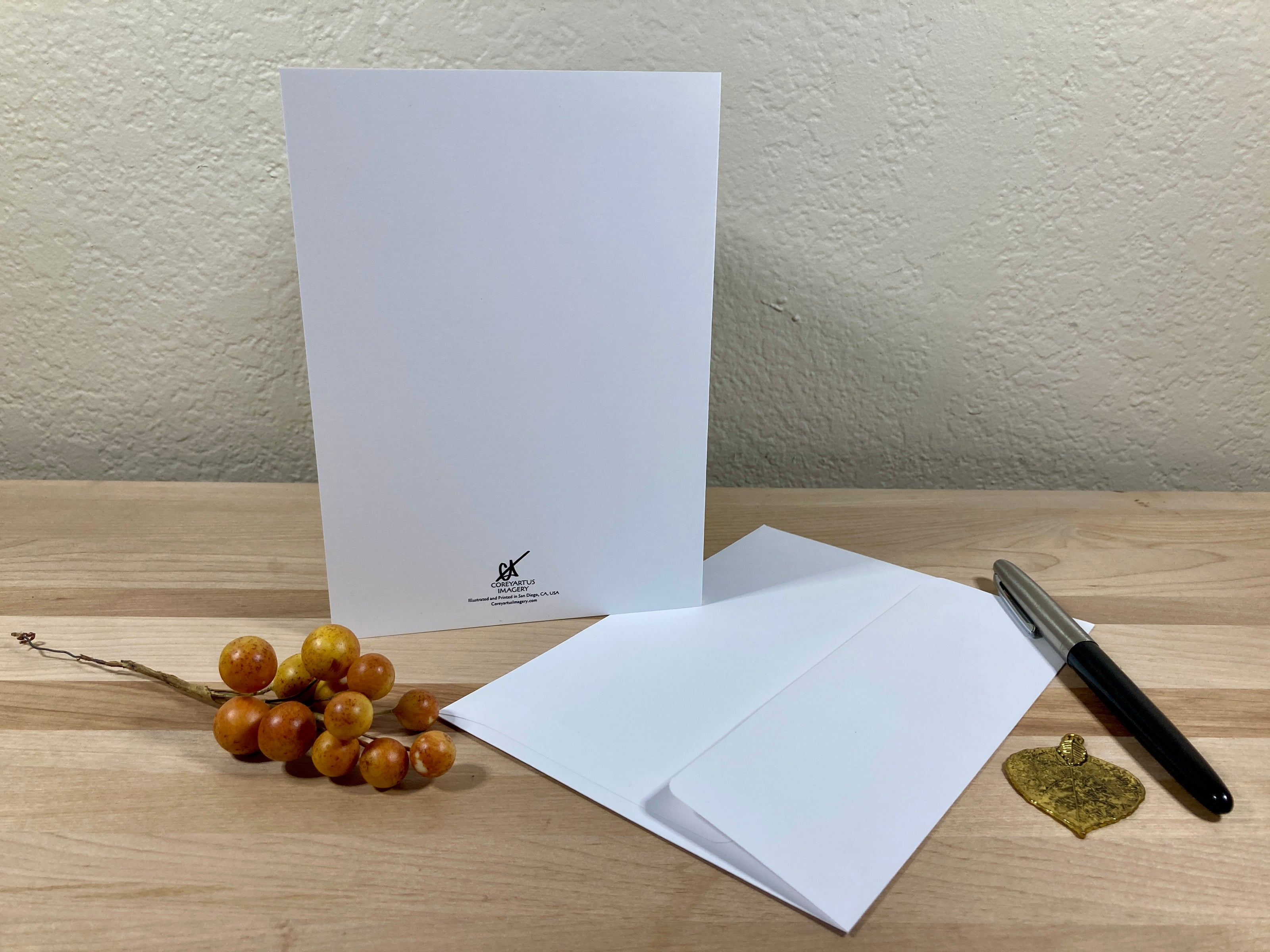 White greeting card, envelope, pen, and decorative items on a wooden surface with a neutral background.