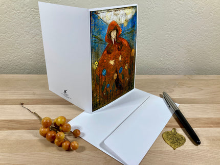 Greeting card with a stylized illustration of a person in a red hood on a wooden surface with a pen and decorative elements.