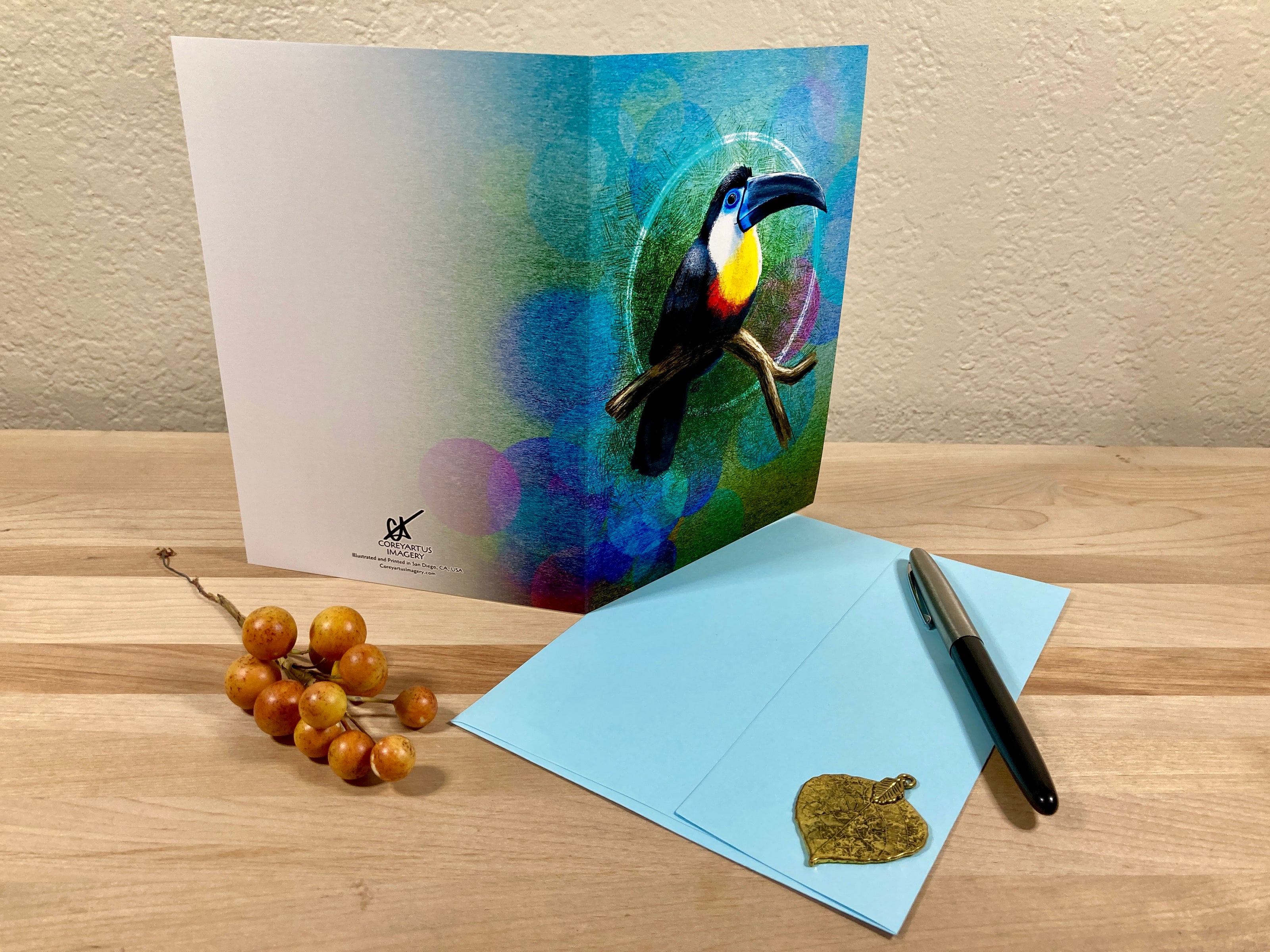 Greeting card with a colorful toucan design on a wooden surface with an envelope and pen.