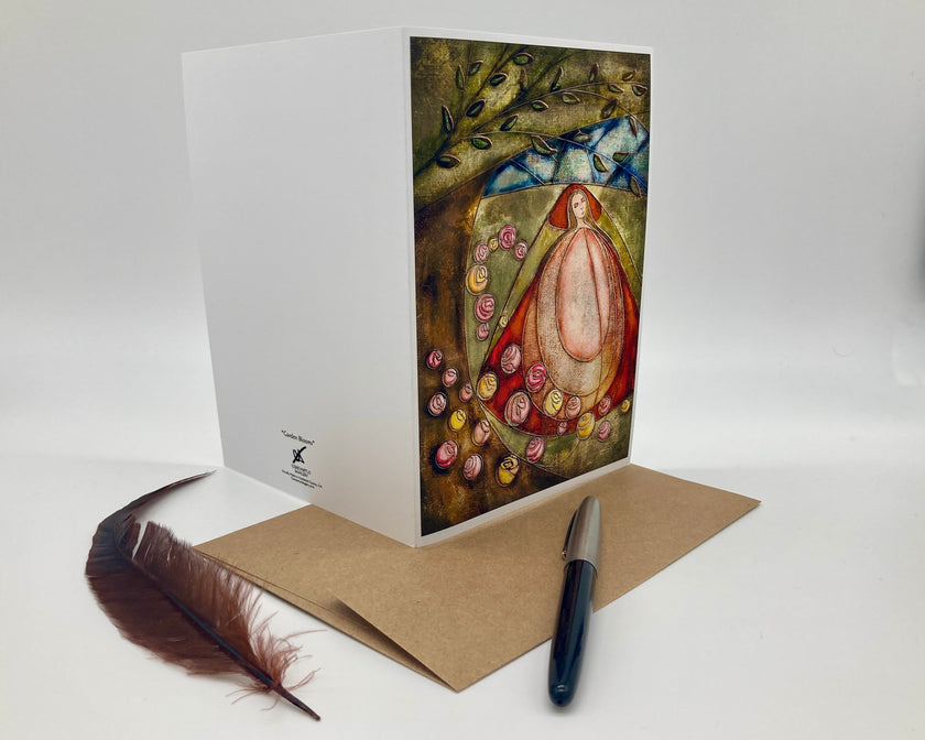 Greeting card with artistic design of a woman surrounded by flowers and leaves, placed on a white surface with a pen and feather.
