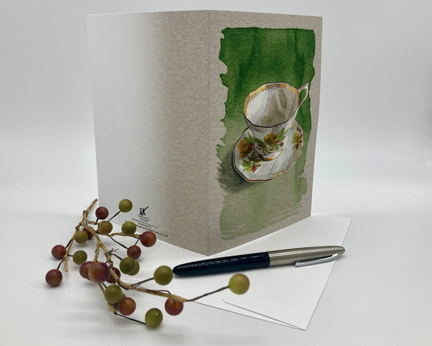Greeting card with a teacup design on a white surface with a pen and decorative branch.