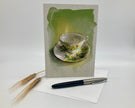 Greeting card with a teacup design on a white surface with a pen and wheat stalks.
