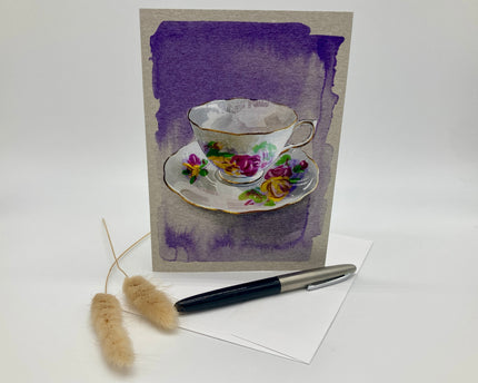 Greeting card with a floral teacup design on a purple watercolor background, accompanied by a pen and decorative elements.