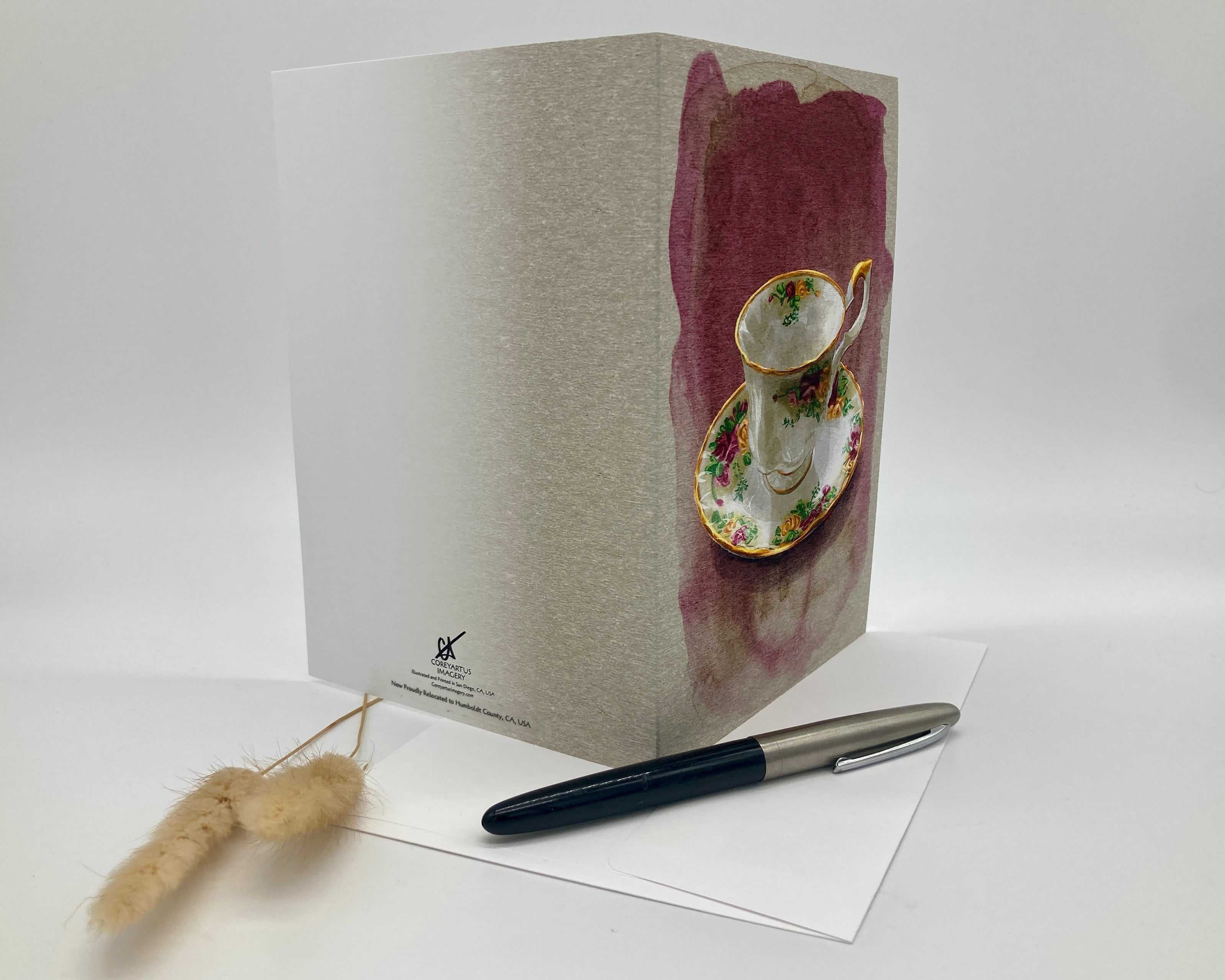 Greeting card with teacup design on a white surface with a pen and decorative item.