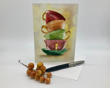 Stack of colorful teacups on a greeting card with a pen and grapes on a white surface