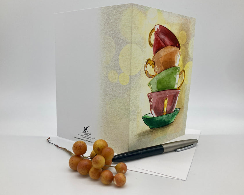 Greeting card with stacked colorful teacups design, pen, and fruit on a white background
